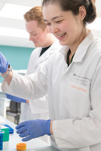 A woman stands in the foreground wearing a white lab coat holding some lab equipment