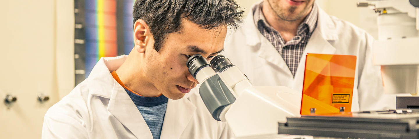 One man looks into a microscope while another man stands behind him