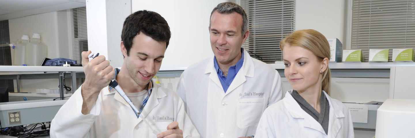 Three people in lab coats stand in a lab looking at a test tube