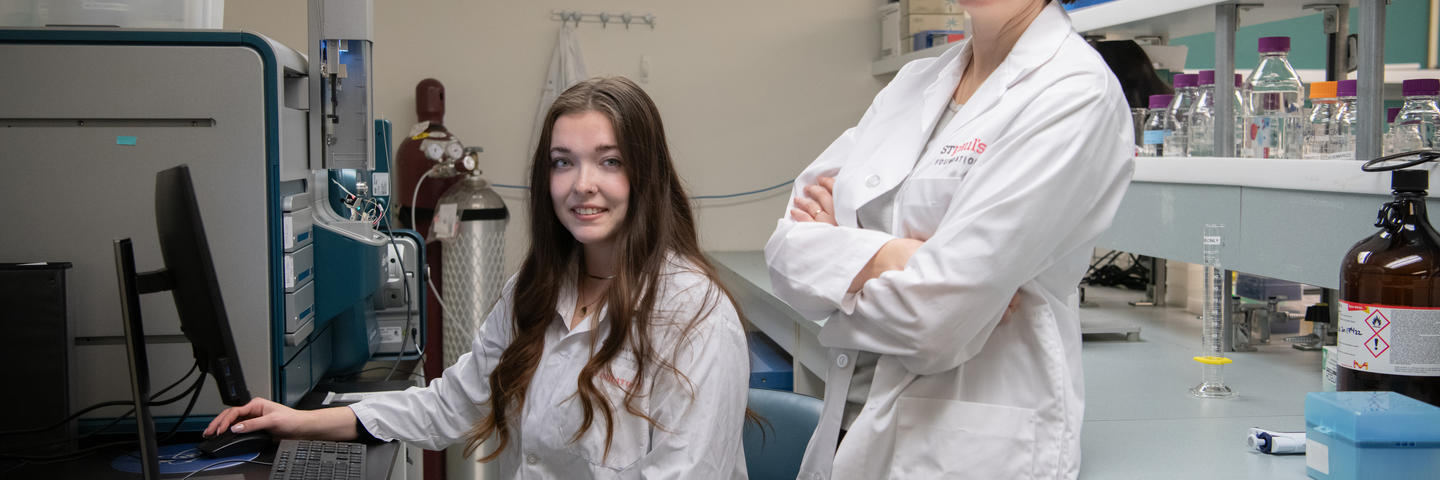 Two women, one seated at a desk and one standing with her arms crossed, are in a lab setting wearing white lab coats