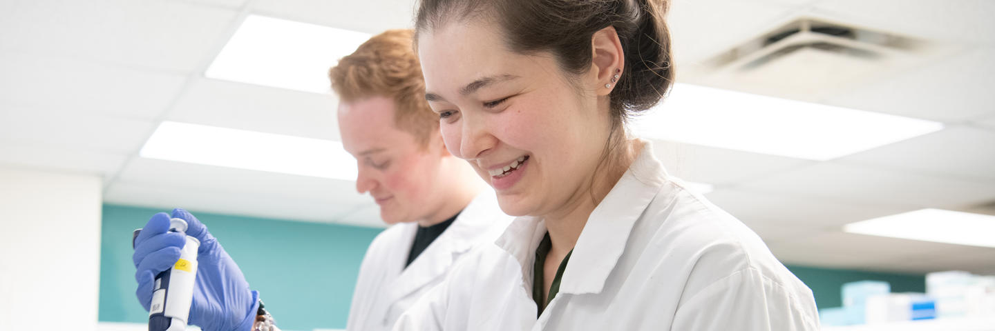 A woman stands in the foreground wearing a white lab coat holding some lab equipment