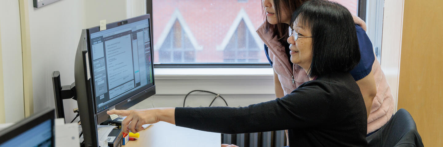 One woman stands behind another woman seated at a desk, pointing to a computer screen